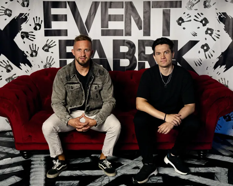 Zwei Männer sitzen auf einem roten Sofa vor einer Wand mit 'EVENT FABRK' in großen Buchstaben. Der Mann auf der linken Seite trägt eine Jeansjacke und weiße Hosen. Der Mann auf der rechten Seite trägt ein schwarzes Hemd und schwarze Hosen.