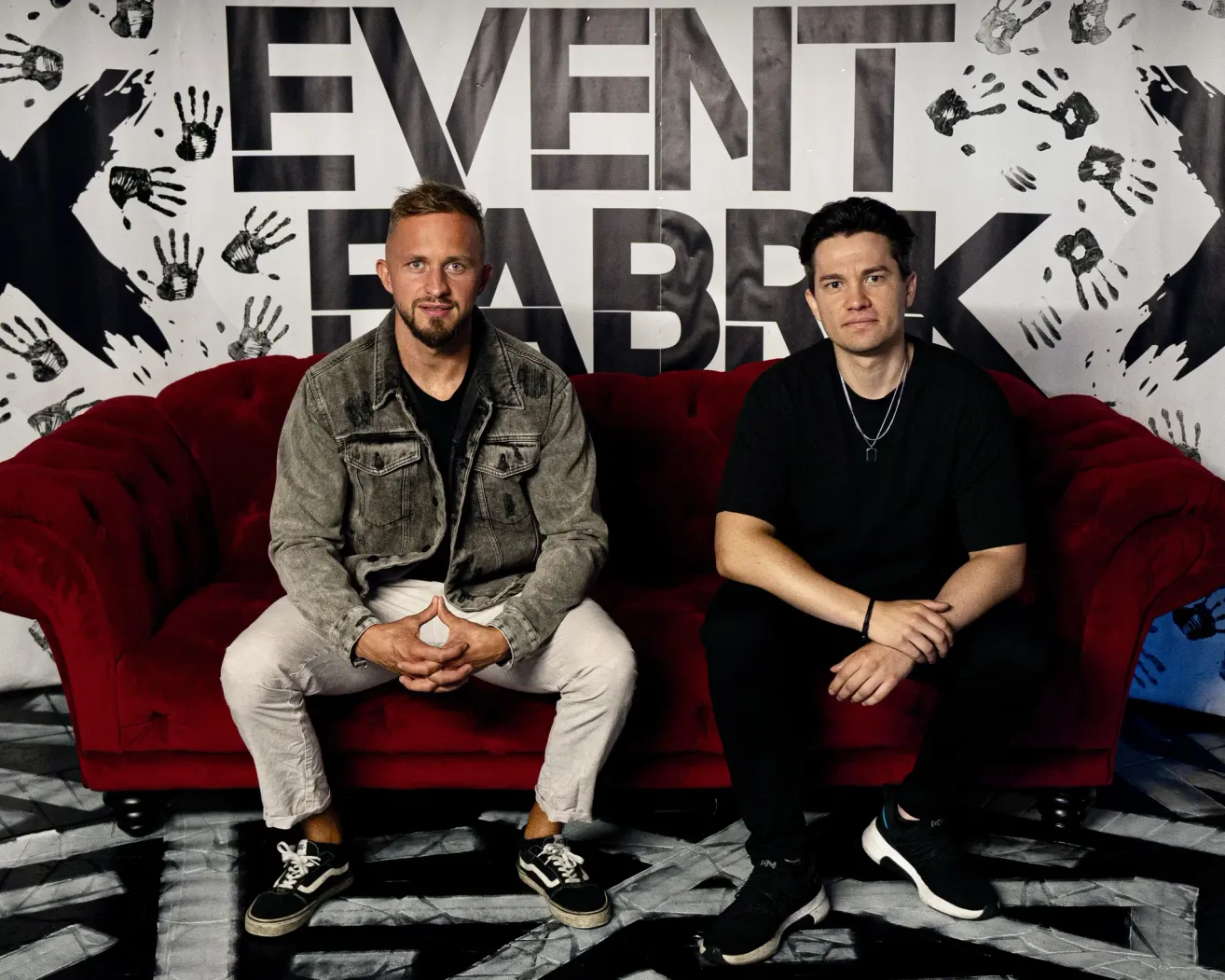 Zwei Männer sitzen auf einem roten Sofa vor einer Wand mit 'EVENT FABRK' in großen Buchstaben. Der Mann auf der linken Seite trägt eine Jeansjacke und weiße Hosen. Der Mann auf der rechten Seite trägt ein schwarzes Hemd und schwarze Hosen.