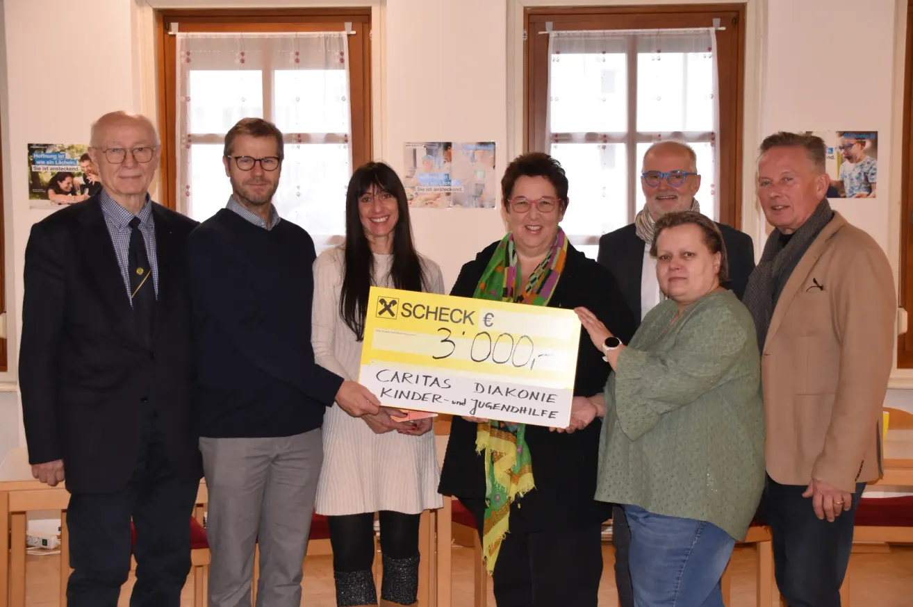 Eine Gruppe von Menschen steht zusammen, hält einen großen Scheck, lächelt und schaut in die Kamera. Der Scheck ist gelb mit einem roten X und sagt 'Caritas Kinder-und Jugendhilfe'.