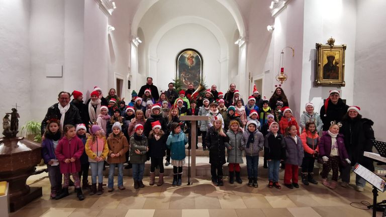 Eine Gruppe von Kindern in Winterkleidung posiert für ein Foto in einer Kirche. Erwachsene und Musikinstrumente sind ebenfalls anwesend.