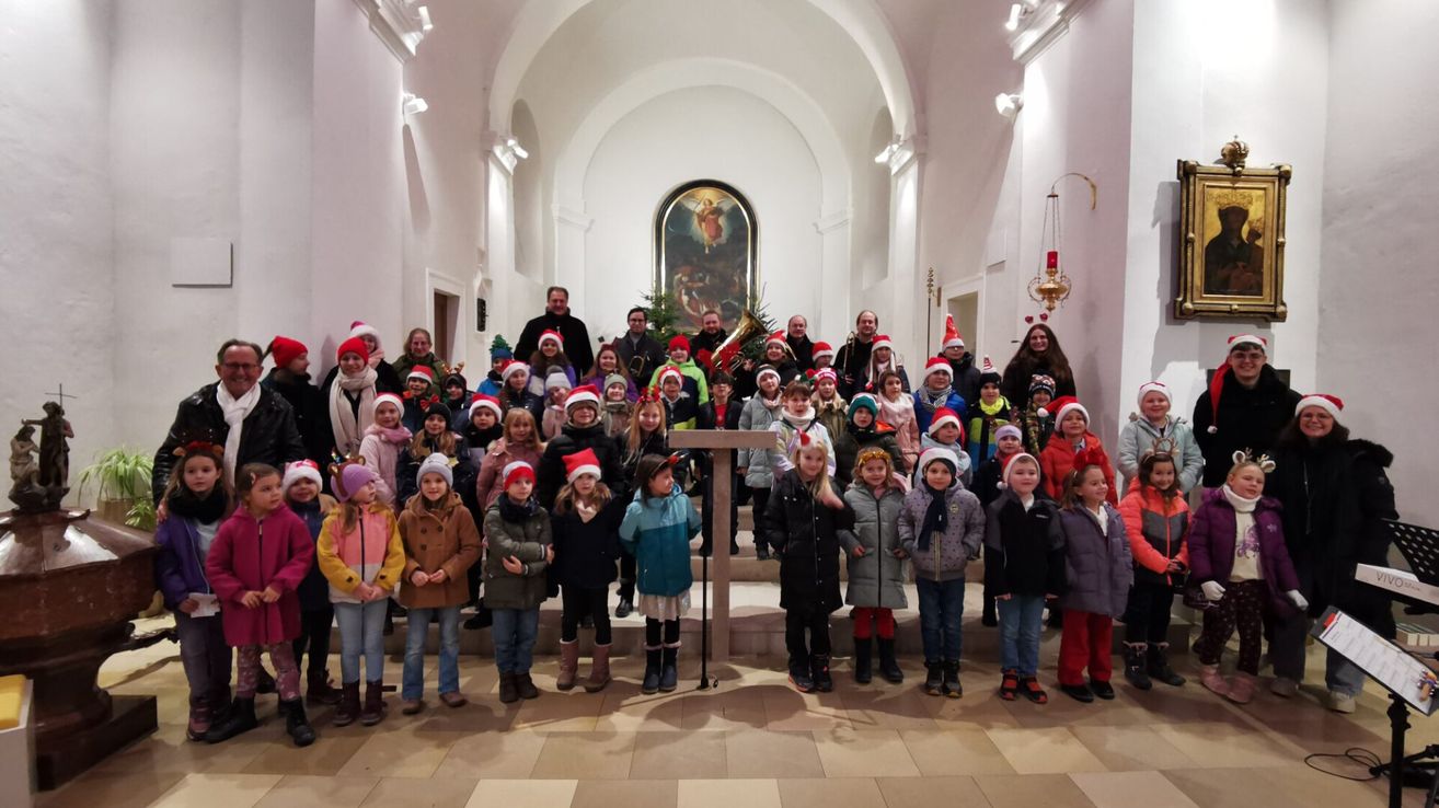 Eine Gruppe von Kindern in Winterkleidung posiert für ein Foto in einer Kirche. Erwachsene und Musikinstrumente sind ebenfalls anwesend.