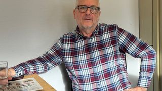 Ein älterer Mann mit Brille und kariertem Hemd sitzt auf einem Stuhl, die Hand auf der Hüfte, mit einer Zeitung auf dem Tisch neben ihm.