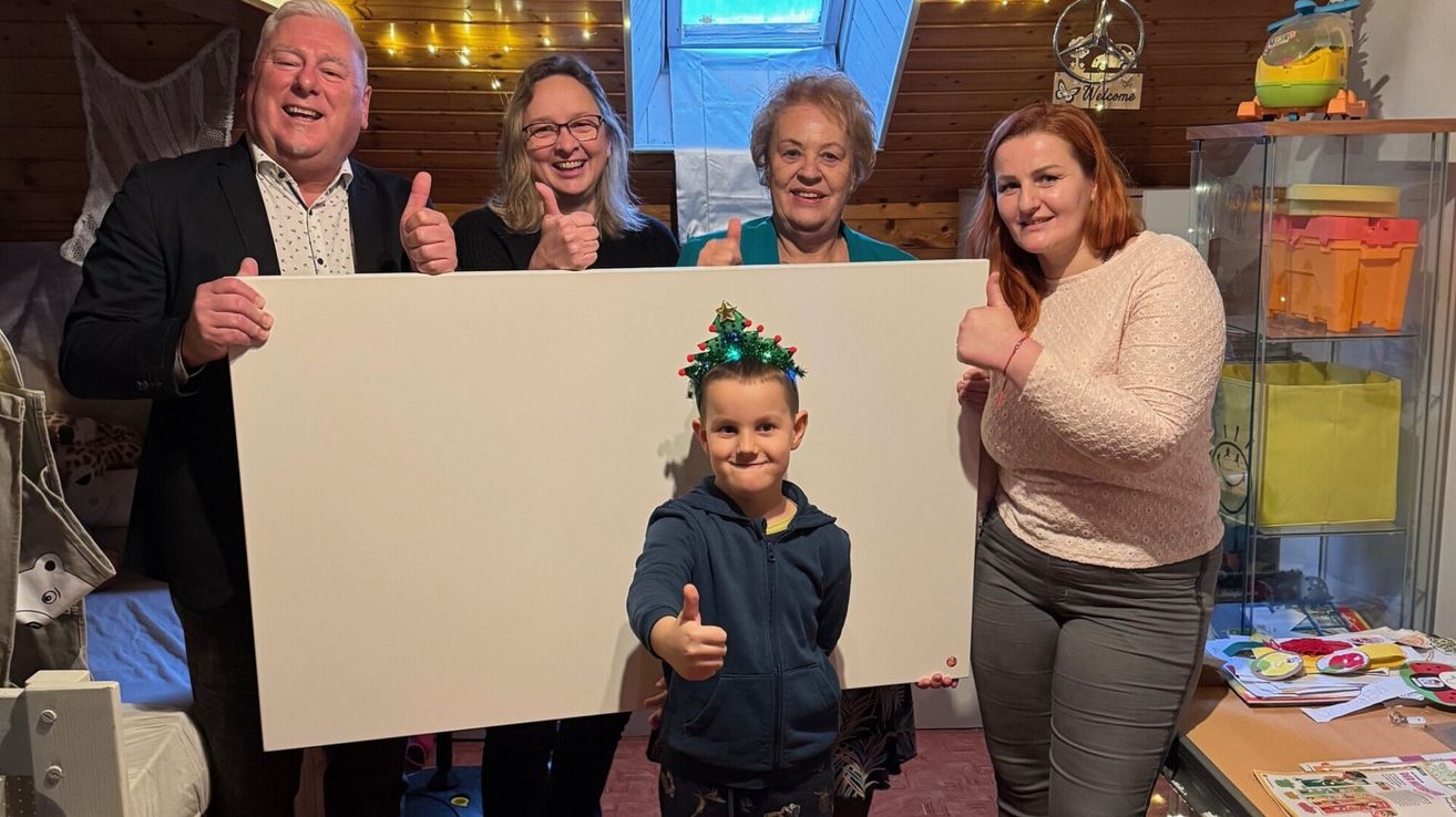 Eine Gruppe von Menschen, darunter ein erwachsener Mann, eine Frau und ein Kind, halten ein großes weißes Brett mit Weihnachtsdekoration. Sie lächeln und geben Daumen hoch.