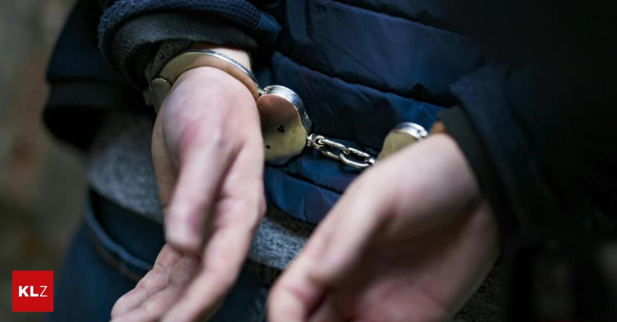 Nahaufnahme der Hände einer Person, die eine blaue Jacke trägt, mit Handschellen an den Handgelenken, vor verwischter Hintergrund.