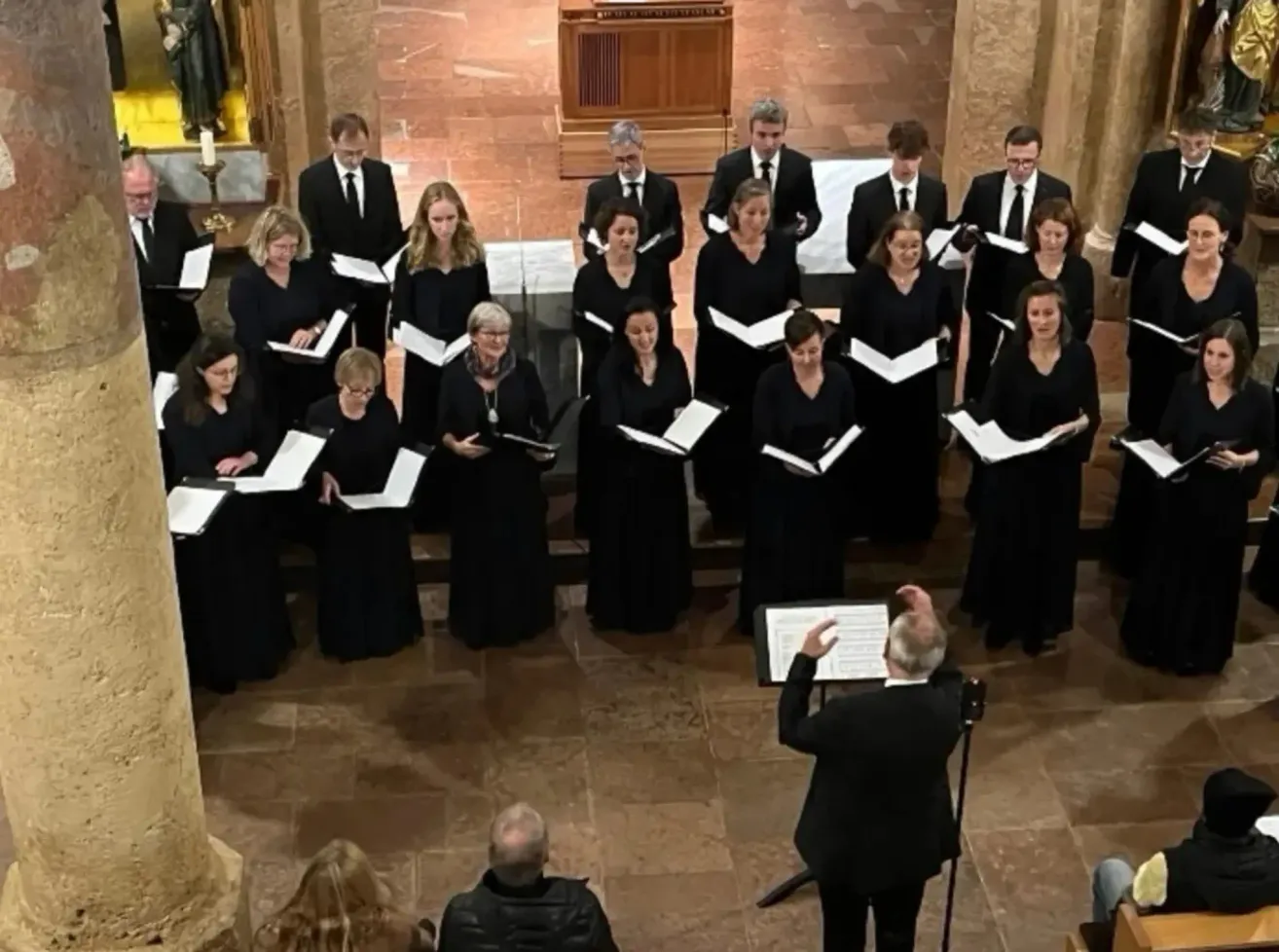 Ein Chor in schwarzer Kleidung, der in einer Kirche mit gefliesten Böden, einer hölzernen Kanzel und Kerzen auftritt, begleitet von einem Dirigenten mit Notenständer.