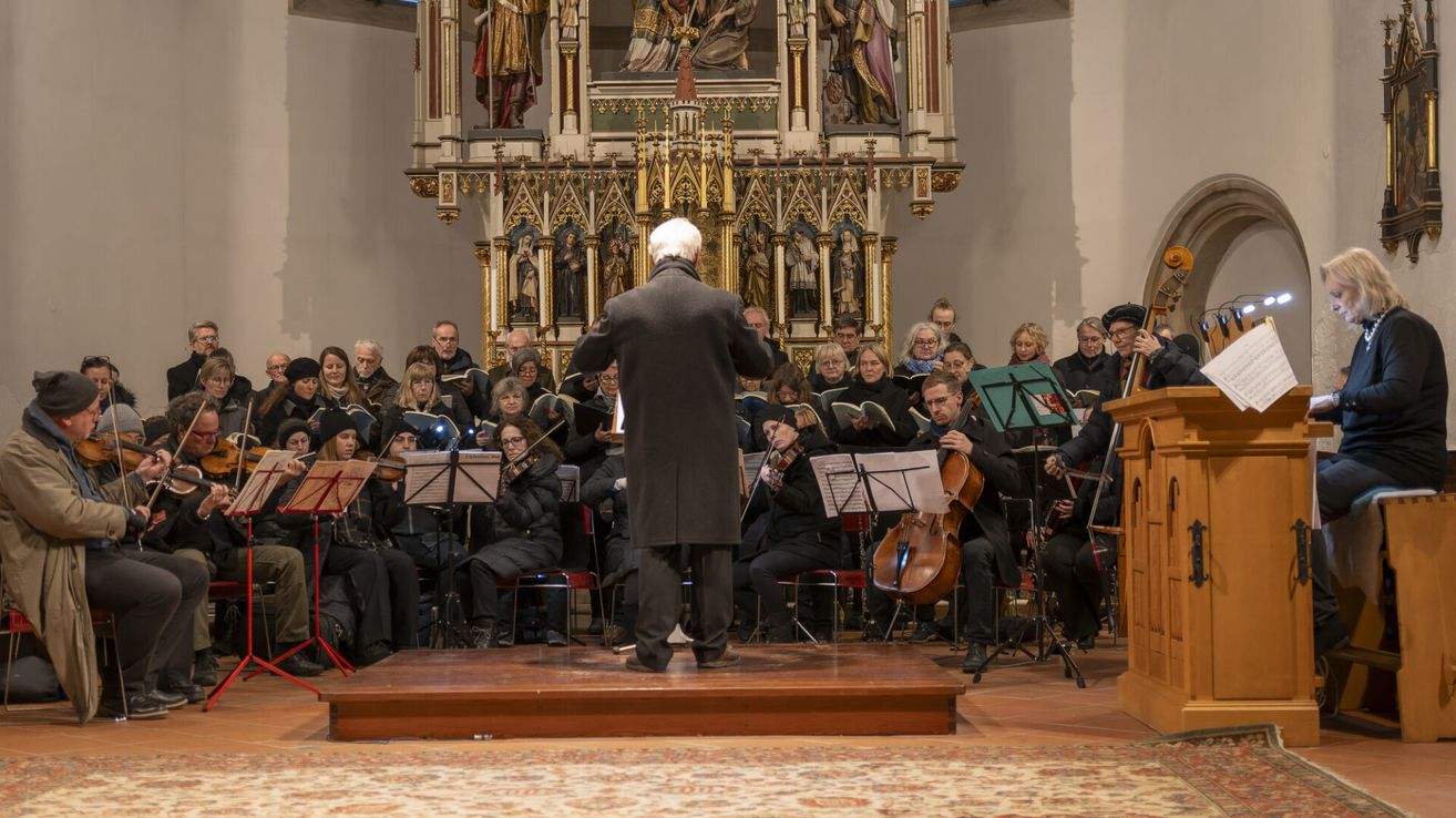 Ein Dirigent steht auf der Bühne und leitet einen Chor und ein Orchester in einer Kirche mit kunstvollen Dekorationen.