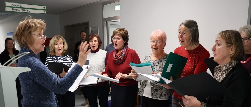 Eine Gruppe älterer Frauen, die Notenblätter halten, singt gemeinsam in einem Raum. Eine Frau vorne gestikuliert mit ihrer Hand. Der Raum hat weiße Wände und eine offene Tür.