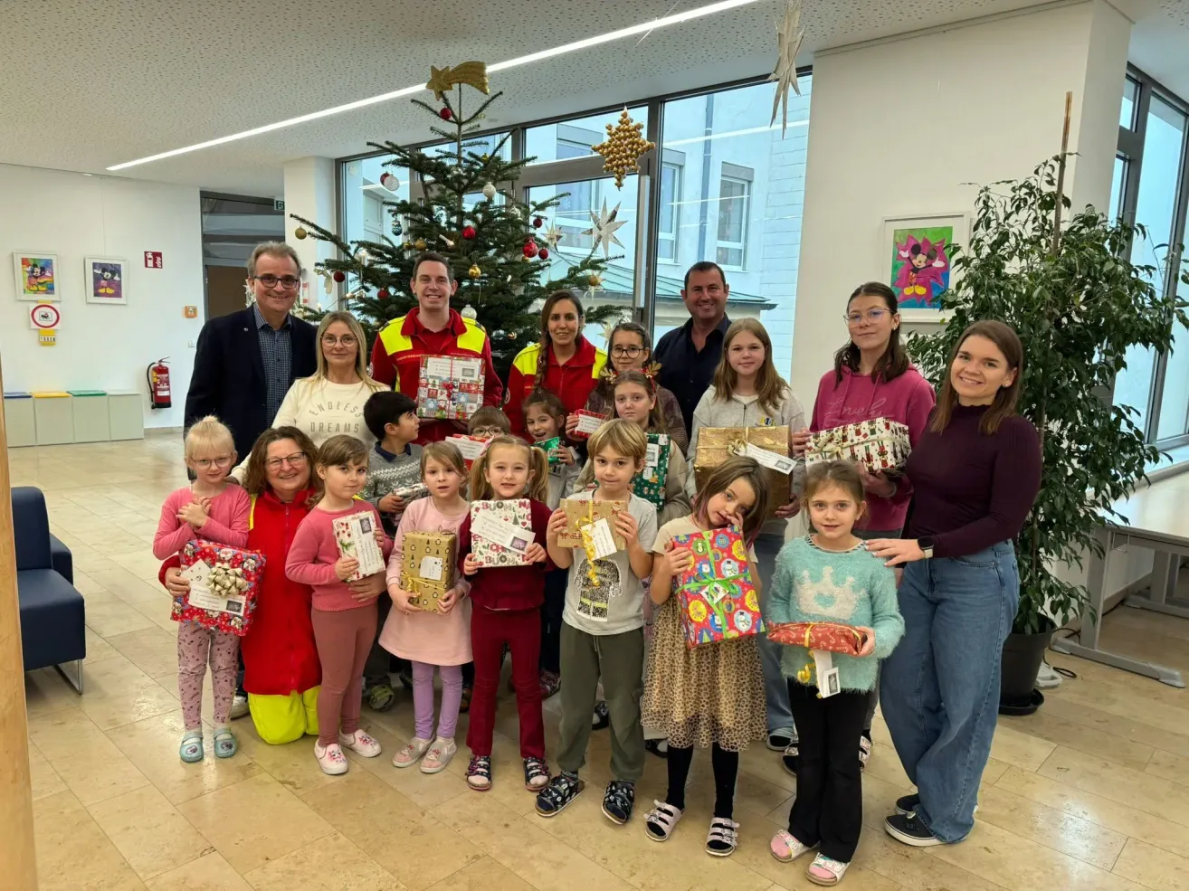 Eine Gruppe von Erwachsenen und Kindern versammelt sich um einen Weihnachtsbaum, hält Geschenke in den Händen und posiert für ein Foto. Dahinter bietet ein Fenster einen Blick auf das Freie.