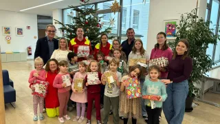 Eine Gruppe von Erwachsenen und Kindern versammelt sich um einen Weihnachtsbaum, hält Geschenke in den Händen und posiert für ein Foto. Dahinter bietet ein Fenster einen Blick auf das Freie.
