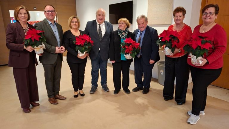 Sechs Personen, feierlich gekleidet, stehen zusammen im Innenbereich. Jede hält eine rote Weihnachtssternpflanze. Ein Mülleimer und ein Fernseher sind im Hintergrund zu sehen.