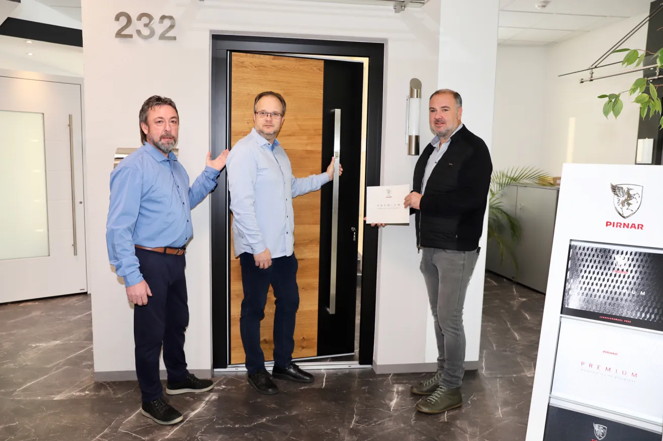 Drei Männer stehen vor einer Tür, einer hält ein Zertifikat. Sie befinden sich in einem modernen Büroraum mit Marmorboden.