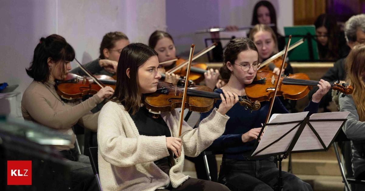 Eine Gruppe junger Frauen spielt Geigen während einer musikalischen Aufführung, wobei eine Geigerin im Fokus ihr Instrument und den Bogen hält, während andere und ein Dirigent im Hintergrund verschwommen sind.