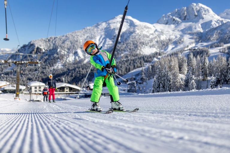Ein junger Skifahrer in einem leuchtend grünen Outfit und mit einer Skibrille fährt den verschneiten Hang hinunter, mit Bergen im Hintergrund.