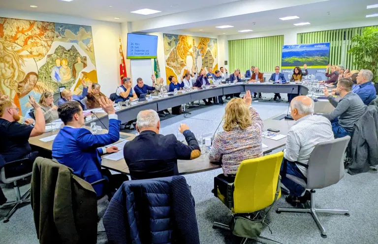 Eine Besprechung findet statt, bei der Menschen die Hände heben, darunter ein Mann und eine Frau. Eine Beamerleinwand zeigt einen Kalender. Mikrofone und Gläser stehen auf dem Tisch.