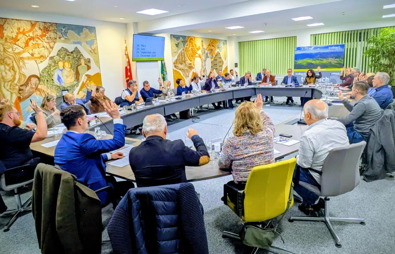 Eine Besprechung findet statt, bei der Menschen die Hände heben, darunter ein Mann und eine Frau. Eine Beamerleinwand zeigt einen Kalender. Mikrofone und Gläser stehen auf dem Tisch.