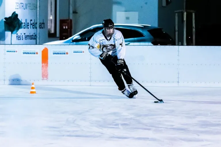 Ein Eishockeyspieler gleitet auf dem Eis und manövriert geschickt mit einem Hockeyschläger, zeigt Beweglichkeit und Präzision. Dahinter zeigt eine Wand Logos, und ein Auto ist geparkt.