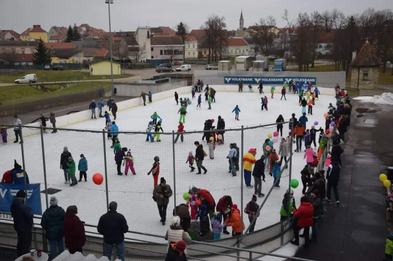 Eine Menschenmenge skat auf einer Eisbahn im Freien mit einer Absperrung. Viele tragen Winterkleidung, und einige halten Luftballons. Im Hintergrund befinden sich Gebäude und Bäume.