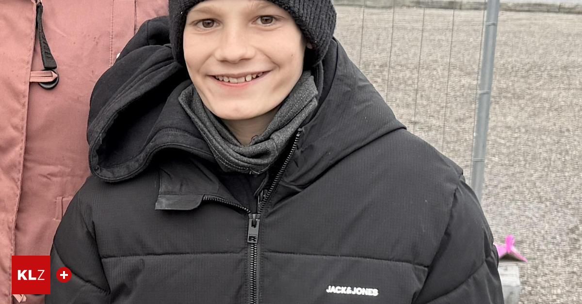 Ein Junge in einer schwarzen Jacke und Mütze, lächelnd, mit einem Schal um den Hals, steht draußen.