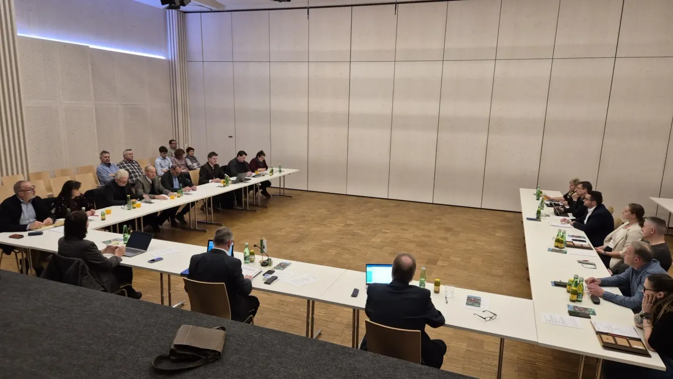 Ein Konferenzraum mit einem langen Tisch. Viele Menschen sitzen auf Stühlen, dem Tisch zugewandt. Einige arbeiten an Laptops. Auf dem Tisch stehen Flaschen und andere Gegenstände.