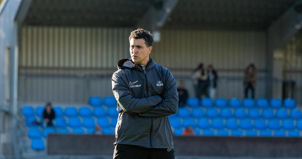 Ein Mann in einer schwarzen Jacke mit den Buchstaben 'FCP' steht auf einem Feld, Arme verschränkt und blickt nach vorne. Hinter ihm sind blaue Tribünen leer. In der Ferne stehen ein paar Leute in der Nähe der Tribünen.