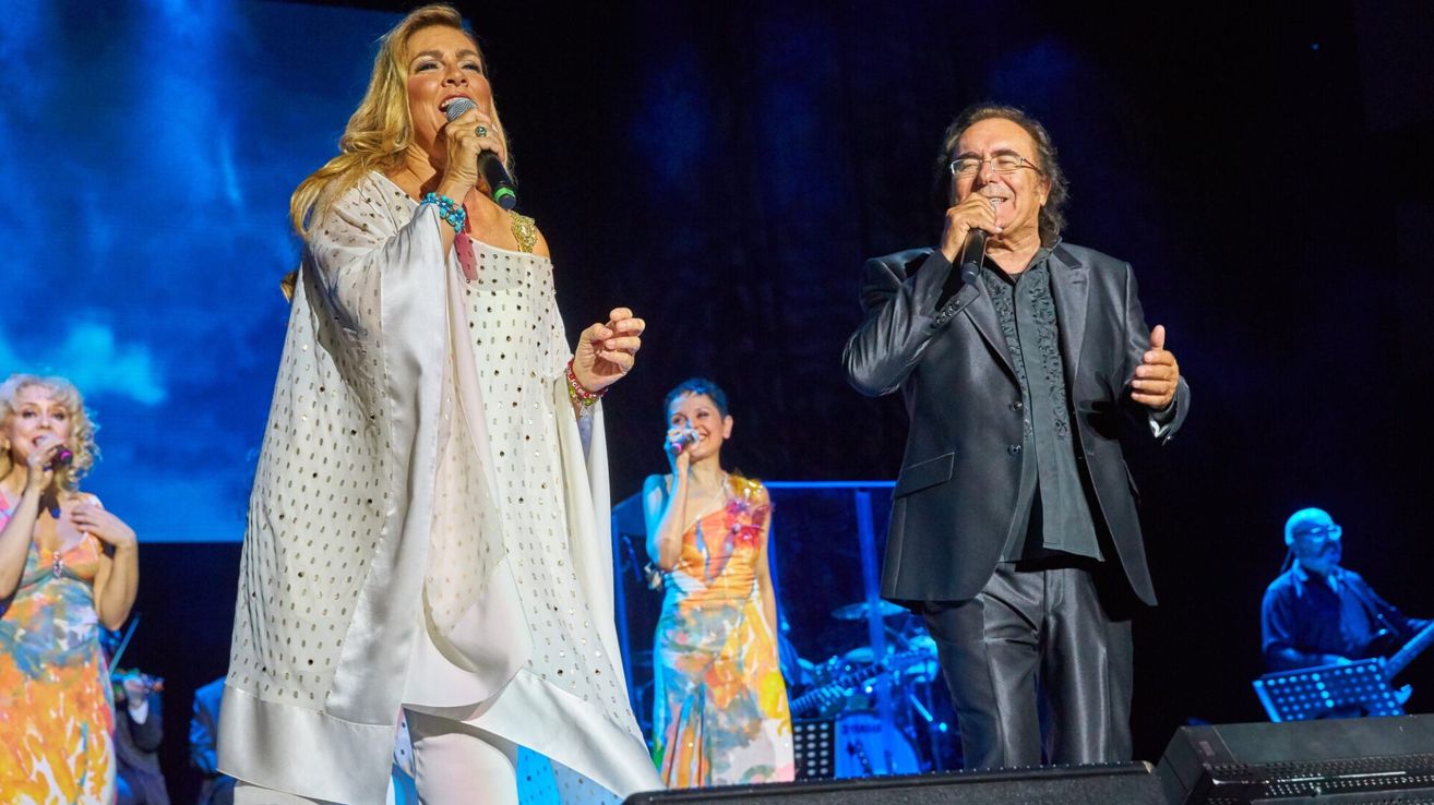 Zwei Künstler stehen auf der Bühne, einer singt in ein Mikrofon, ein anderer neben ihr, und eine Frau in einem farbenfrohen Kleid singt im Hintergrund.