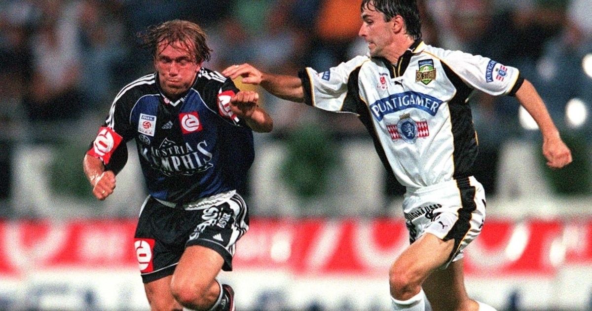 Zwei männliche Fußballspieler konkurrieren in einem Spiel. Einer trägt ein blaues Trikot mit 'Austria', der andere ein weißes Trikot mit 'Puntigamer'.