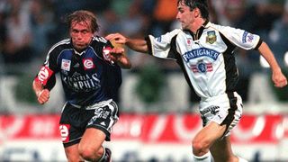 Zwei männliche Fußballspieler konkurrieren in einem Spiel. Einer trägt ein blaues Trikot mit 'Austria', der andere ein weißes Trikot mit 'Puntigamer'.