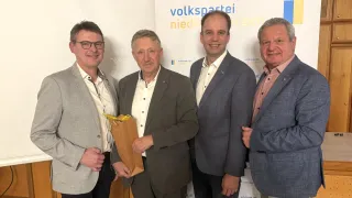 Vier Männer stehen zusammen vor einer Wand mit dem Logo der Volkspartei Niederoesterreich. Ein Mann hält einen braunen Papierbeutel.