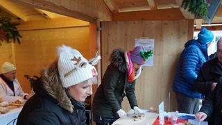 Mehrere Menschen versammeln sich in einer Holzkabine und bereiten Essen zu. Eine Frau in einer Hirschmütze schneidet Essen auf einem Schneidebrett. Eine andere Frau schreibt auf eine Tafel. Zwei Männer beobachten.