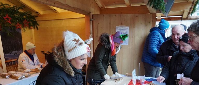 Mehrere Menschen versammeln sich in einer Holzkabine und bereiten Essen zu. Eine Frau in einer Hirschmütze schneidet Essen auf einem Schneidebrett. Eine andere Frau schreibt auf eine Tafel. Zwei Männer beobachten.