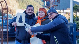 Three people are standing in an outdoor area, smiling and posing for a photo. A man on the left is holding a board with a tree logo. A woman in the middle is wearing a scarf. The man on the right is holding a bottle and a box. Behind them is a mountain with trees.