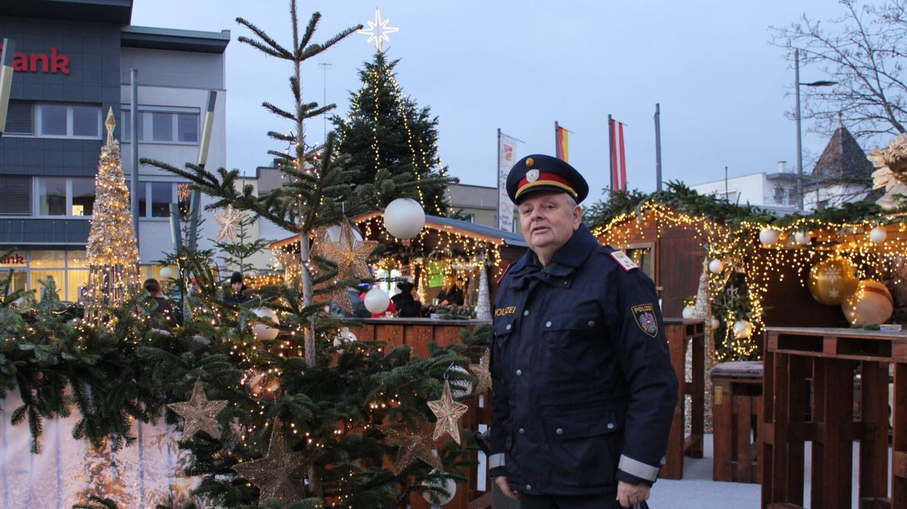Ein Polizeibeamter steht vor einem geschmückten Weihnachtsbaum mit Lichtern und Kugeln. Im Hintergrund befinden sich weitere geschmückte Bäume und eine festliche Atmosphäre mit Lichtern.