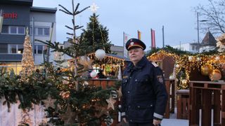 Ein Polizeibeamter steht vor einem geschmückten Weihnachtsbaum mit Lichtern und Kugeln. Im Hintergrund befinden sich weitere geschmückte Bäume und eine festliche Atmosphäre mit Lichtern.