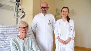 Drei Menschen stehen in einem Krankenhauszimmer. Eine lächelnde ältere Frau sitzt in einem Krankenhausbett. Zwei Menschen in weißen Laborkittel stehen neben ihr.