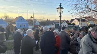 Eine Gruppe von Menschen, wahrscheinlich Senioren, versammelte sich draußen in einem Dorf, in der Nähe von Gebäuden und Straßenlaternen, an einem sonnigen Tag.