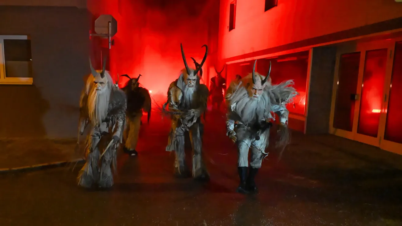 Eine Gruppe von Menschen in Hörnerkostümen geht in einer Reihe, beleuchtet von rotem Licht. Sie sind vor einem Gebäude mit einer roten Wand.