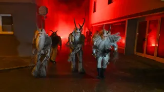 Eine Gruppe von Menschen in Hörnerkostümen geht in einer Reihe, beleuchtet von rotem Licht. Sie sind vor einem Gebäude mit einer roten Wand.