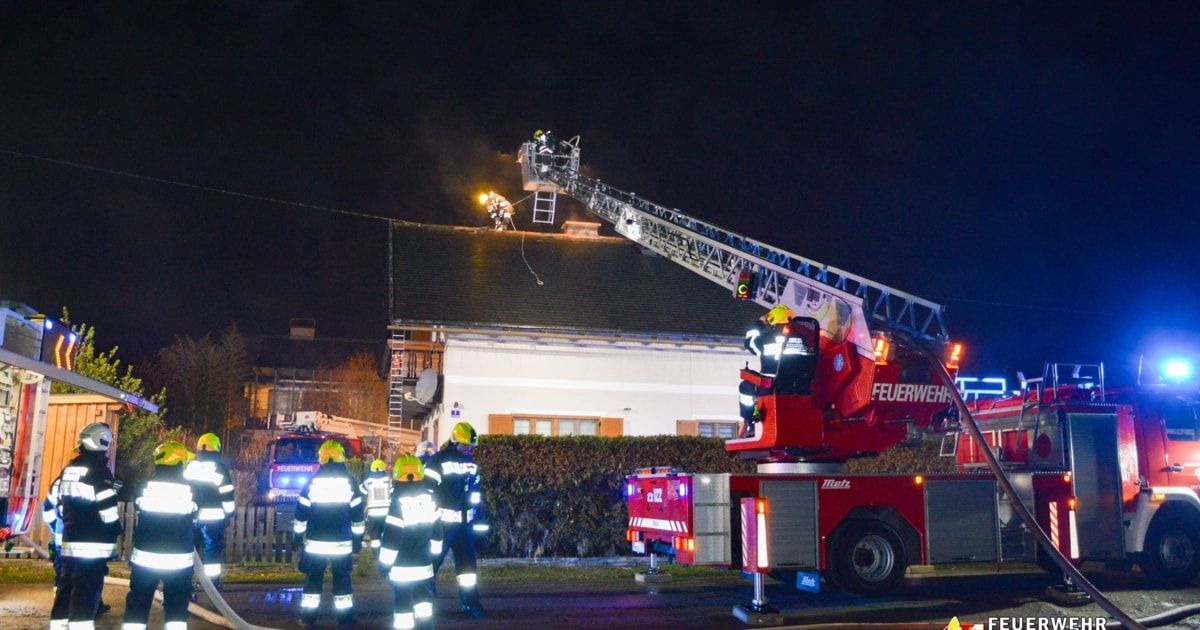 Feuerwehrleute in Schutzkleidung sind am Brandort eines Hauses, mit einem Feuerwehrwagen und einer Leiter in Position.