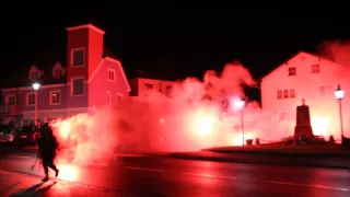 Eine Straße in der Nacht wird von rotem Licht, möglicherweise von einem Feuer, beleuchtet. Rauch erfüllt die Luft, und ein Gebäude ist im Hintergrund sichtbar.