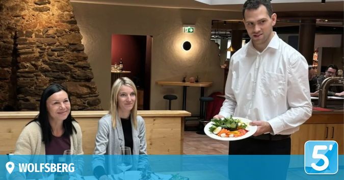 Ein Kellner in einem weißen Hemd hält einen Teller mit Gemüse in einem Restaurant. Zwei Frauen sitzen vor ihm an einem Tisch.