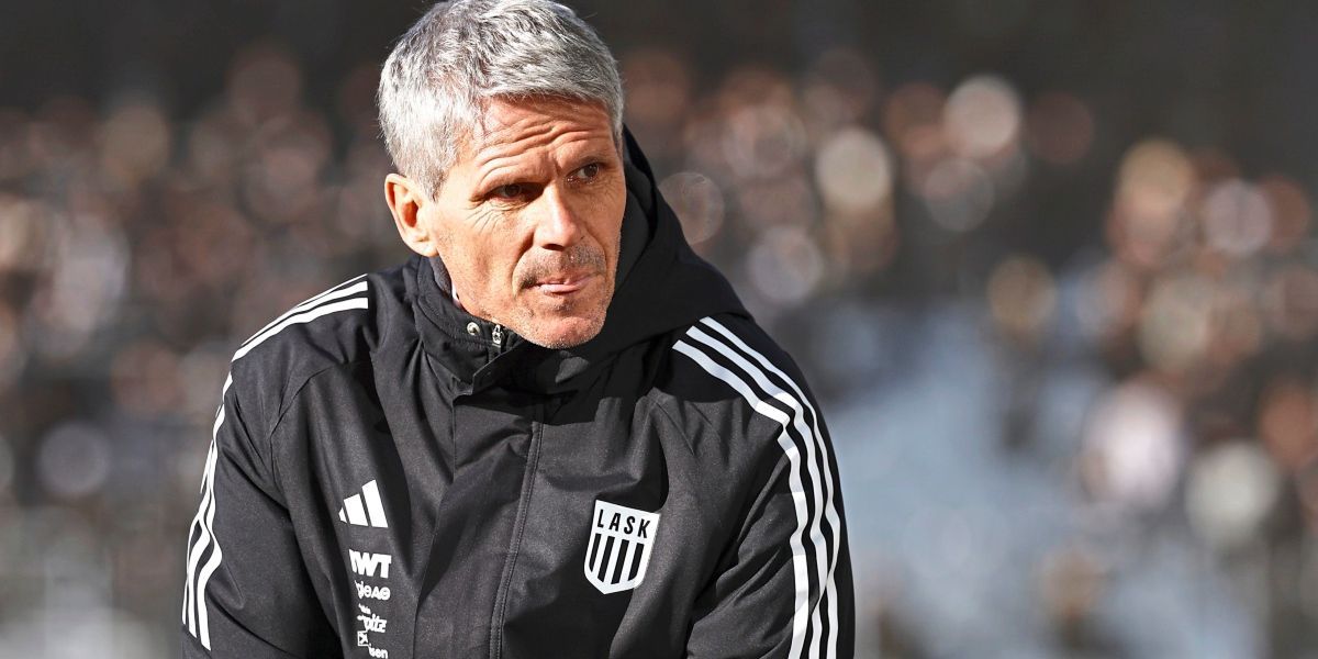 Ein älterer Mann in einem schwarzen Jacke mit weißen Streifen und einer Kapuze steht auf einem Feld und sieht ernst aus.