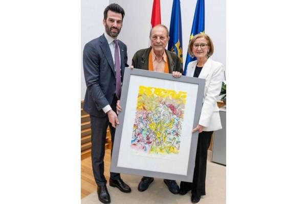 Three people stand holding a framed painting, with two men dressed in suits and the woman in formal attire. Behind them, flags are displayed on the wall.