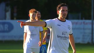 A soccer player in a white uniform with the word 'Genussfleisch' on the front is pointing to the left, possibly giving instructions during a game. Two other players in white jerseys are in the background.