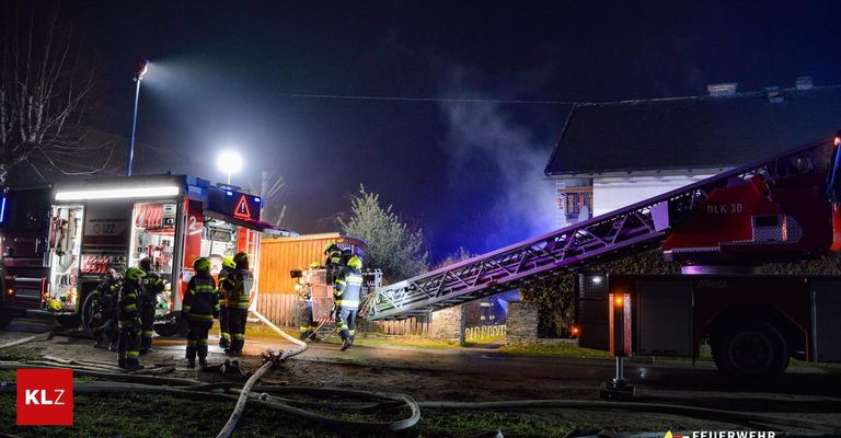 Feuerwehrleute reagieren nachts auf einen Hausbrand. Sie benutzen eine Leiter, um das Dach zu erreichen. Rauch kommt aus dem Haus.