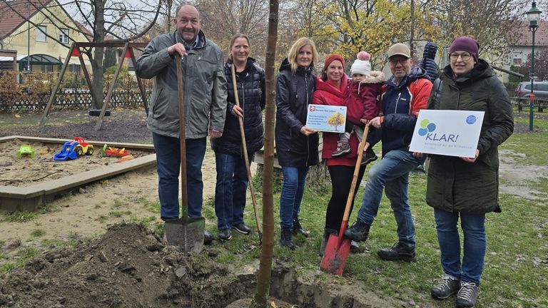 Eine Gruppe von Erwachsenen und Kindern posiert mit Schaufeln um einen frisch gepflanzten Baum in einem Park.