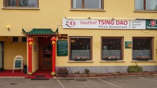 Ein gelbes Gebäude mit einem Schild, das Gasthof TSING DAO China Restaurant liest. Der Eingang hat einen roten Teppich und rote Pfosten mit chinesischen Laternen. Es gibt drei Fenster mit Topfpflanzen und Vorhängen.