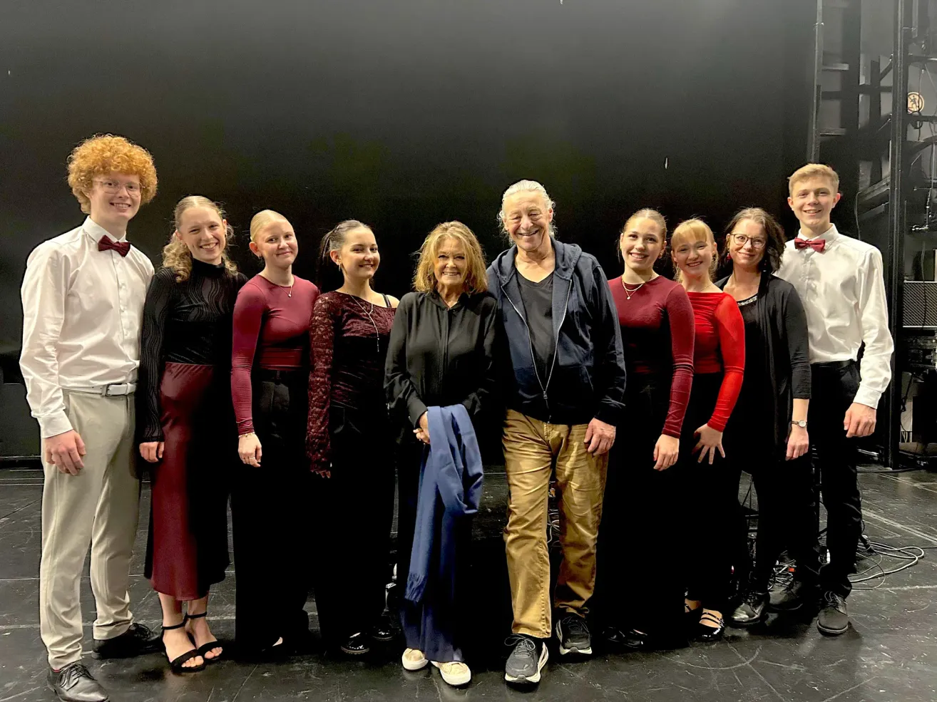 Eine Gruppe junger Frauen und Männer steht mit einer lächelnden älteren Frau und einem Mann auf der Bühne. Sie tragen alle formelle Kleidung.