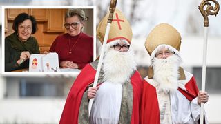 Zwei Personen in Bischofskostümen, eine mit einem Stab, posieren draußen. Eine Frau in einem roten Pullover hält ein Papier mit einer Zeichnung.