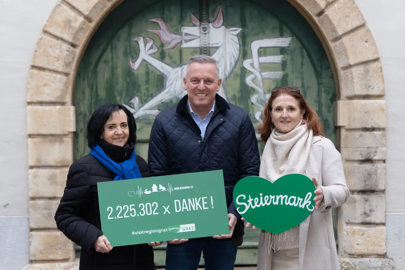 Drei Personen posieren mit Plakaten vor einer kunstvollen grünen Tür. Der Mann hält ein herzförmiges Schild mit der Aufschrift 'Stiermark'. Die Frau links hält ein grünes Schild mit der Nummer 2.225.302 und der Botschaft 'x Danke!'. Die Frau rechts trägt einen Schal. Hinter ihnen ist eine Wandmalerei eines weißen Einhorns.