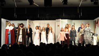 Eine Gruppe von Menschen tritt auf einer Bühne auf. Die Bühne hat weiße Wände und Türen. Einige Menschen tragen weiße Kleider, andere tragen Jacken.